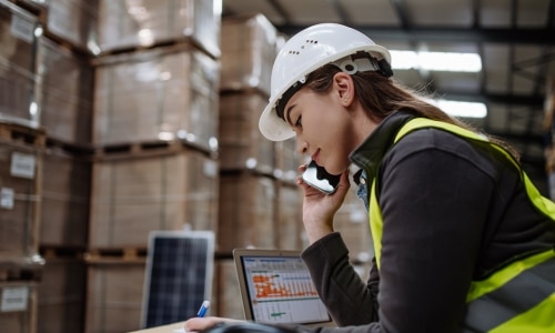 Kobieta w kasku rozmawiająca przez telefon na magazynie WMS